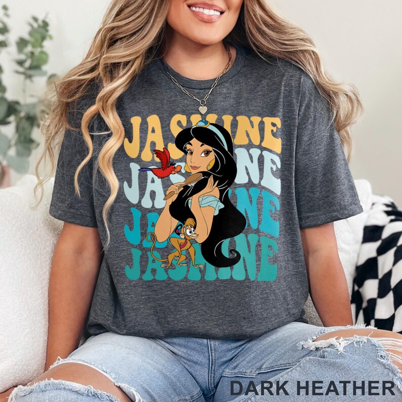 May include: Dark heather gray t-shirt featuring a graphic of Princess Jasmine with the text "Jasmine" in a retro font. The design includes a parrot and a monkey. The shirt is made of a soft material.