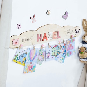 May include: A wooden cloud-shaped display with the name "HAZEL" in pink letters. It features a string with small pegs holding children's artwork. Decorative wooden shapes of a pig, bunny, flowers, and a butterfly are also present.