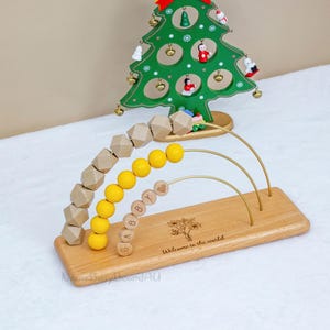 Puede incluir: Juguete de &aacute;baco de madera con cuentas amarillas y de color natural, y el nombre "Gabby" escrito. La base est&aacute; grabada con un &aacute;rbol y las palabras "Welcome to the world". Una decoraci&oacute;n de &aacute;rbol de Navidad verde est&aacute; en el fondo.