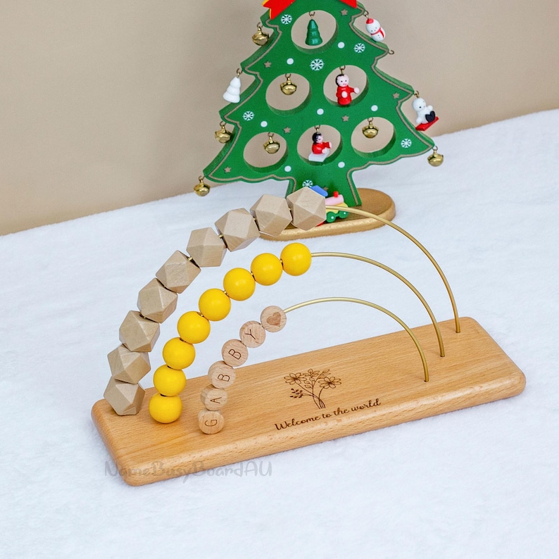 Puede incluir: Juguete de &aacute;baco de madera con cuentas amarillas y de color natural, y el nombre "Gabby" escrito. La base est&aacute; grabada con un &aacute;rbol y las palabras "Welcome to the world". Una decoraci&oacute;n de &aacute;rbol de Navidad verde est&aacute; en el fondo.
