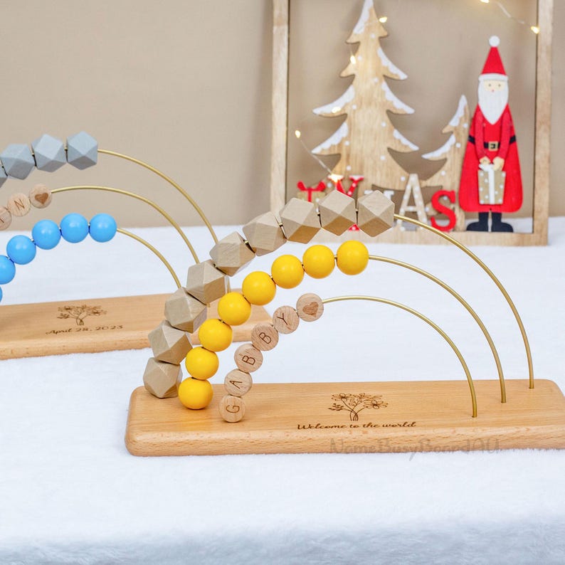 Puede incluir: &Aacute;baco de madera con cuentas de colores y bloques de nombres personalizados. El &aacute;baco presenta una base de madera con arcos de alambre dorados. Las cuentas son en tonos grises, azules y amarillos. La base est&aacute; grabada con las palabras "Bienvenido al mundo."