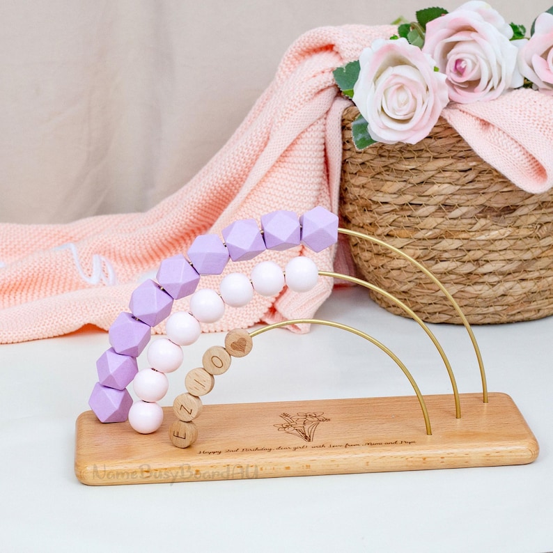 Puede incluir: &Aacute;baco de beb&eacute; de madera con base de madera clara, con tres alambres met&aacute;licos arqueados. El &aacute;baco tiene filas de formas geom&eacute;tricas lavanda, esferas blancas y cuentas de madera que deletrean "EZIO". La base est&aacute; grabada con texto y un dise&ntilde;o floral.