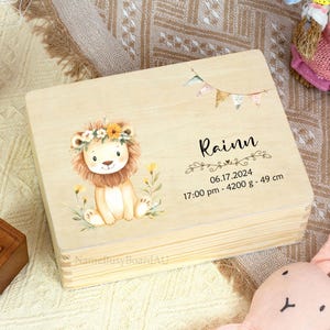 May include: A light-coloured wooden box featuring a watercolour illustration of a lion cub wearing a floral crown. The box is personalised with the name "Rainn", date, time, weight (4200 g), and height (49 cm). A small banner with pastel flags is also featured.