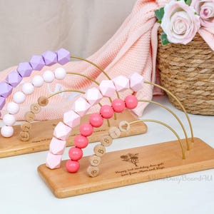 Puede incluir: Juguete de &aacute;baco de madera con cuentas rosas, moradas y blancas. Las cuentas est&aacute;n dispuestas en arcos de alambre dorado. La base de madera est&aacute; grabada con texto y un dise&ntilde;o floral. El texto dice "Happy 2nd Birthday, dear girls. Love from Mimi and Papi."