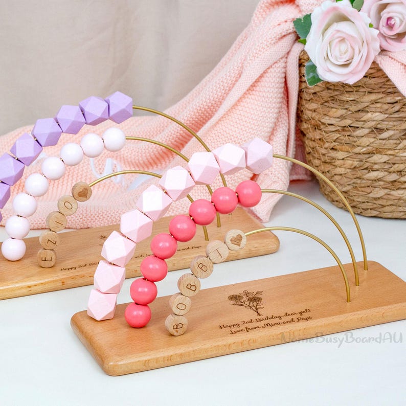 Puede incluir: Juguete de &aacute;baco de madera con cuentas rosas, moradas y blancas. Las cuentas est&aacute;n dispuestas en arcos de alambre dorado. La base de madera est&aacute; grabada con texto y un dise&ntilde;o floral. El texto dice "Happy 2nd Birthday, dear girls. Love from Mimi and Papi."
