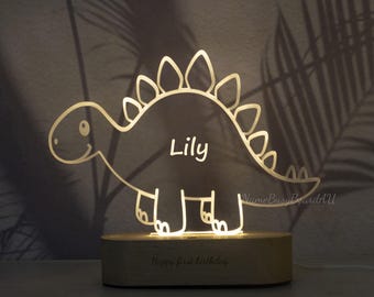 Puede incluir: Luz de noche con forma de dinosaurio brillante, con el nombre "Lily" en blanco. La lámpara está sobre una base de madera con la inscripción "Happy first birthday". Un objeto decorativo para la habitación de los niños.