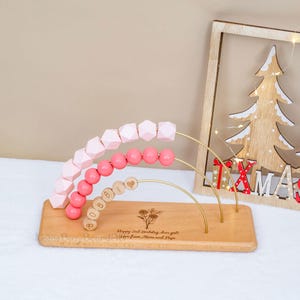Puede incluir: &Aacute;baco de madera con cuentas rosas y formas geom&eacute;tricas. La base est&aacute; grabada con texto y un dise&ntilde;o floral. Una decoraci&oacute;n de &aacute;rbol de Navidad de madera est&aacute; en el fondo.