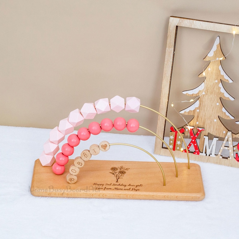 Puede incluir: &Aacute;baco de madera con cuentas rosas y formas geom&eacute;tricas. La base est&aacute; grabada con texto y un dise&ntilde;o floral. Una decoraci&oacute;n de &aacute;rbol de Navidad de madera est&aacute; en el fondo.