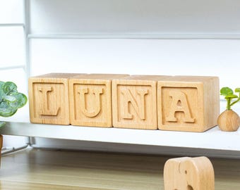 May include: Wooden blocks spelling "LUNA" in capital letters. The blocks are light brown and sit on a white shelf. A small wooden elephant figurine is in front of the blocks. A small plant is to the left of the blocks.
