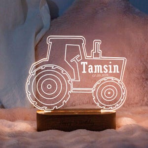 May include: A tractor-shaped LED night light with the name "Tamsin" and the date "07.09.2024" in white. The wooden base has the inscription "Happy 1st Birthday". The light is designed for a child's room.