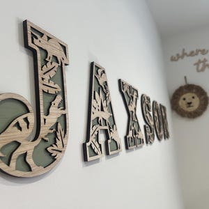 May include: Wooden wall letters spelling "JAXSON" with a dinosaur theme. The letters are light brown with a black outline and a green background. The letter "J" features a dinosaur and leaves.