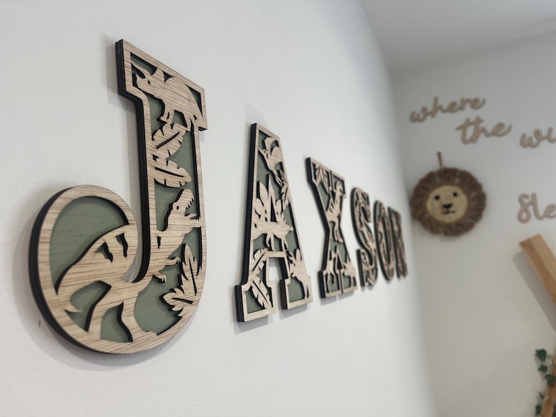 May include: Wooden wall letters spelling "JAXSON" with a dinosaur theme. The letters are light brown with a black outline and a green background. The letter "J" features a dinosaur and leaves.