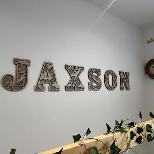 May include: Wooden wall decor spelling out the name "JAXSON" in layered letters. Each letter has a light brown outer layer and a green and brown inner layer with a dinosaur theme. A lion head and the words "where the" are also on the wall.