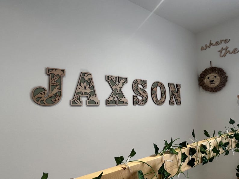May include: Wooden wall decor spelling out the name "JAXSON" in layered letters. Each letter has a light brown outer layer and a green and brown inner layer with a dinosaur theme. A lion head and the words "where the" are also on the wall.