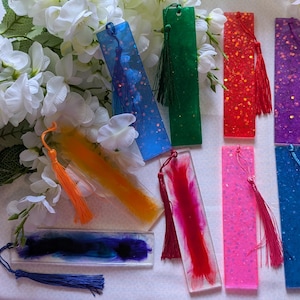 May include: Assortment of eight colourful resin bookmarks with glitter and tassels. The bookmarks are rectangular and feature various colours, including blue, green, red, pink, and orange.