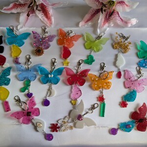 Può includere: Una collezione di portachiavi a farfalla colorati con vari disegni e abbellimenti. I portachiavi presentano glitter, ciondoli e accenti pendenti nelle tonalità di blu, rosa, arancione e verde. Ogni portachiavi misura circa 5-7 cm di lunghezza.