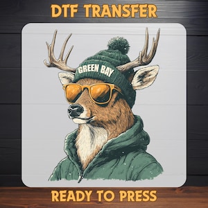May include: A DTF transfer featuring a deer wearing a green beanie with "GREEN BAY" text, orange sunglasses, and a green jacket. The words "DTF TRANSFER" and "READY TO PRESS" are also displayed.
