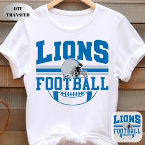 May include: White t-shirt featuring a blue and grey football-themed design. The design includes the word "LIONS" in bold blue letters, a grey football helmet, and the word "FOOTBALL" in blue. A smaller version of the design is in the lower right corner.