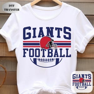 May include: White t-shirt with a navy blue and red design that says "GIANTS FOOTBALL". The design includes a red football helmet and a football graphic. The shirt is hanging on a wooden hanger.