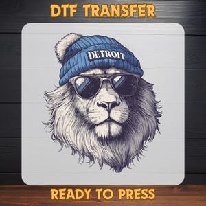 May include: DTF transfer featuring a lion wearing a blue beanie with "DETROIT" and sunglasses. The lion has a white and gray mane. The words "READY TO PRESS" are at the bottom.