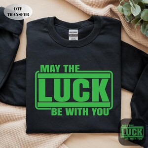 May include: Black sweatshirt with the text "MAY THE LUCK BE WITH YOU" in green. The design is in a rectangular shape. The sweatshirt has a crew neck and long sleeves. The image also includes a small version of the design.