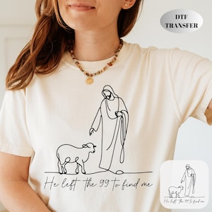 Peut inclure: T-shirt crème avec un dessin au trait noir d'une figure et d'un agneau, avec le texte "He left the 99 to find me." Un transfert DTF est visible. Le t-shirt est à col rond et à manches courtes.