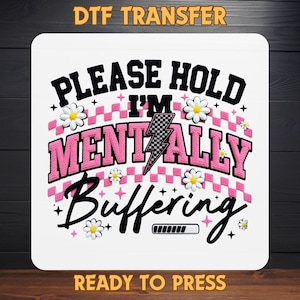 May include: White DTF transfer with the text "PLEASE HOLD I'M MENTALLY BUFFERING" in black and pink. The design includes daisy accents and a lightning bolt. The words "DTF TRANSFER" and "READY TO PRESS" are also present.