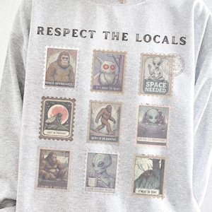 Cozy cryptid sweatshirt with a grid of vintage postage stamp illustrations including Mothman, Bigfoot, and alien folklore characters shown on a model wearing a Gildan 18000 Ash crewneck.