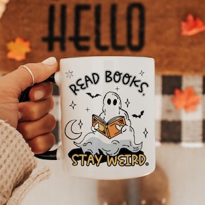May include: A white ceramic mug featuring a black graphic of a ghost reading an orange book, surrounded by stars, a crescent moon, and bats. The text reads "READ BOOKS" at the top and "STAY WEIRD" at the bottom in a bold, black, sans-serif font.