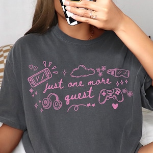 Dark gray cozy gamer aesthetic shirt with pink graphic text just one more quest worn in relaxed indoor setting