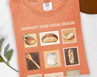 May include: Orange Comfort Colors t-shirt with the text "SUPPORT YOUR LOCAL DEALER" and illustrations of bread and baking tools. The shirt features a crew neck and short sleeves. The design includes images resembling postage stamps.
