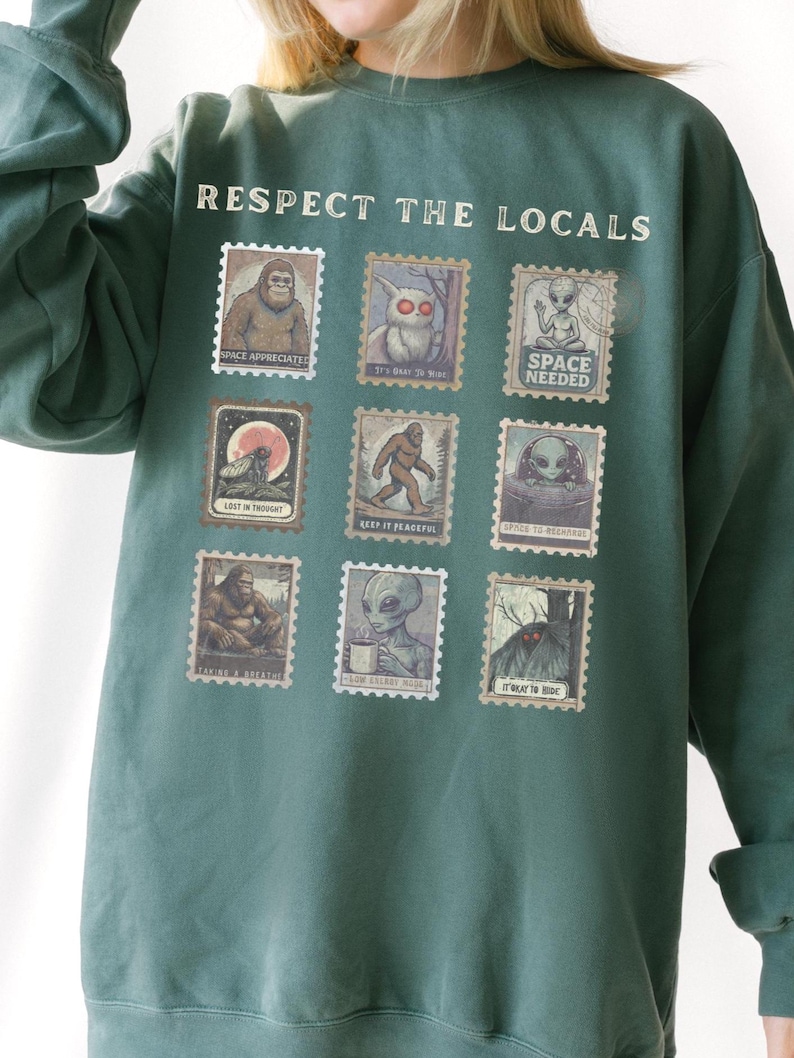 Cozy cryptid sweatshirt with a grid of vintage postage stamp illustrations including Mothman, Bigfoot, and alien folklore characters shown on a model wearing a Comfort Colors 1566 Blue Spruce crewneck.