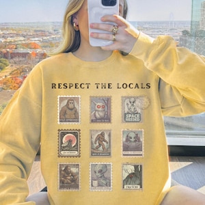 Cozy cryptid sweatshirt with a grid of vintage postage stamp illustrations including Mothman, Bigfoot, and alien folklore characters shown on a model wearing a Comfort Colors 1566 Butter crewneck.