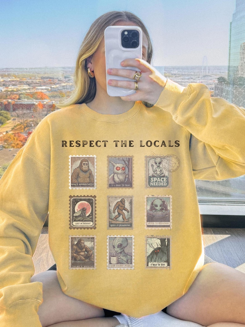 Cozy cryptid sweatshirt with a grid of vintage postage stamp illustrations including Mothman, Bigfoot, and alien folklore characters shown on a model wearing a Comfort Colors 1566 Butter crewneck.