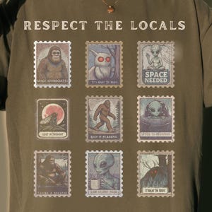 A Pepper colored Comfort Colors t-shirt with "Respect The Locals" cryptid stamp graphic, featuring Bigfoot, Mothman, and Alien stamps. Ideal for cryptid lovers and introverts.