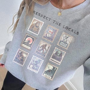 Cozy cryptid sweatshirt with a grid of vintage postage stamp illustrations including Mothman, Bigfoot, and alien folklore characters shown on a model wearing a Gildan 18000 Sport Grey crewneck.