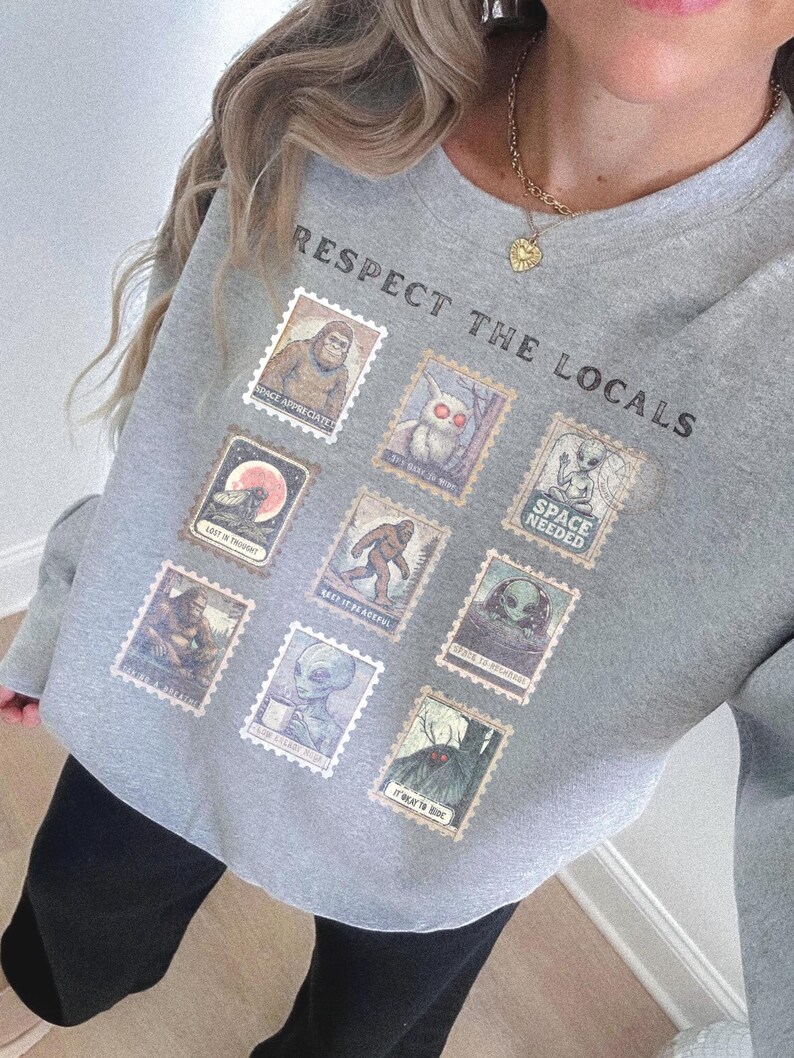 Cozy cryptid sweatshirt with a grid of vintage postage stamp illustrations including Mothman, Bigfoot, and alien folklore characters shown on a model wearing a Gildan 18000 Sport Grey crewneck.