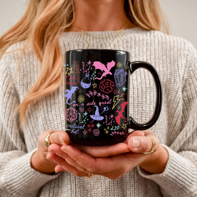 K&ouml;nnte beinhalten: Schwarze Keramiktasse mit farbenfrohen Fantasy-Illustrationen. Die Tasse zeigt einen Drachen, W&uuml;rfel, eine Zaubertrankflasche und Text mit der Aufschrift "on a side quest" und "critical hit". Die Tasse hat einen gro&szlig;en schwarzen Henkel.
