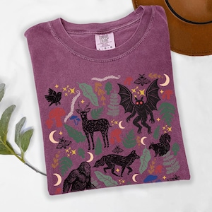 A Berry Comfort Colors t-shirt featuring a forest-themed linocut cryptid design with Mothman, Bigfoot, and forest animals surrounded by mushrooms, ferns, stars, and moons in vintage woodland colors.