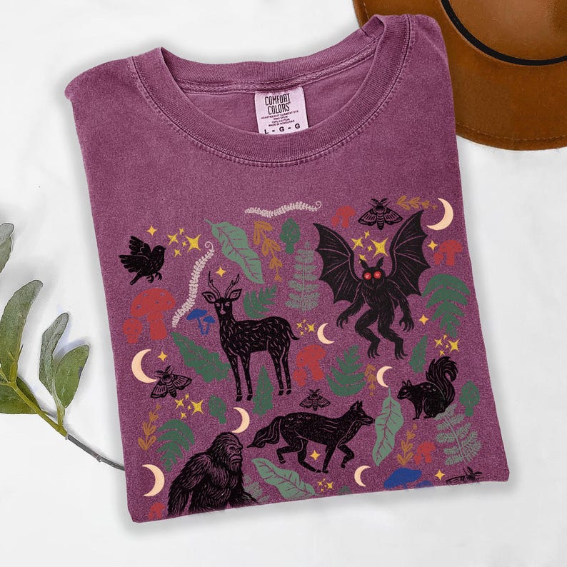 A Berry Comfort Colors t-shirt featuring a forest-themed linocut cryptid design with Mothman, Bigfoot, and forest animals surrounded by mushrooms, ferns, stars, and moons in vintage woodland colors.