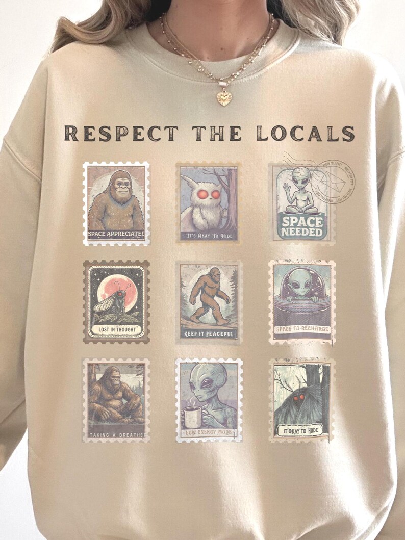 Cozy cryptid sweatshirt with a grid of vintage postage stamp illustrations including Mothman, Bigfoot, and alien folklore characters shown on a model wearing a Gildan 18000 Sand crewneck.