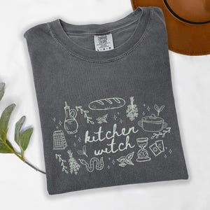 Kitchen Witch Comfort Colors t-shirt in Pepper gray featuring a cottagecore kitchen witch graphic with bread loaf, hanging herbs, cooking pot, olive oil bottle, tarot cards, snake, and botanical accents.