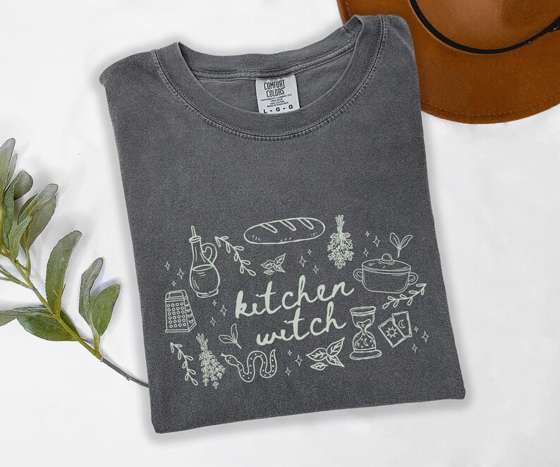 Kitchen Witch Comfort Colors t-shirt in Pepper gray featuring a cottagecore kitchen witch graphic with bread loaf, hanging herbs, cooking pot, olive oil bottle, tarot cards, snake, and botanical accents.