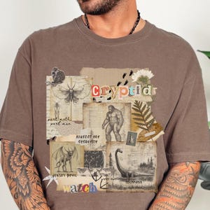 A man wears a Comfort Colors Espresso t shirt with a Cryptozoology design featuring Mothman, Bigfoot, Lochness and the Jersey Devil in a vintage field journal style. Intended for camping, hiking and outdoorsy activities or for lovers of spooky folklore.