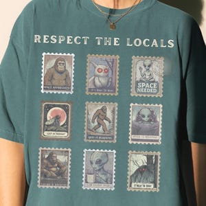 A woman wears a Blue Spruce colored Comfort Colors t-shirt with "Respect The Locals" cryptid stamp graphic, featuring Bigfoot, Mothman, and Alien stamps. Ideal for cryptid lovers and introverts.