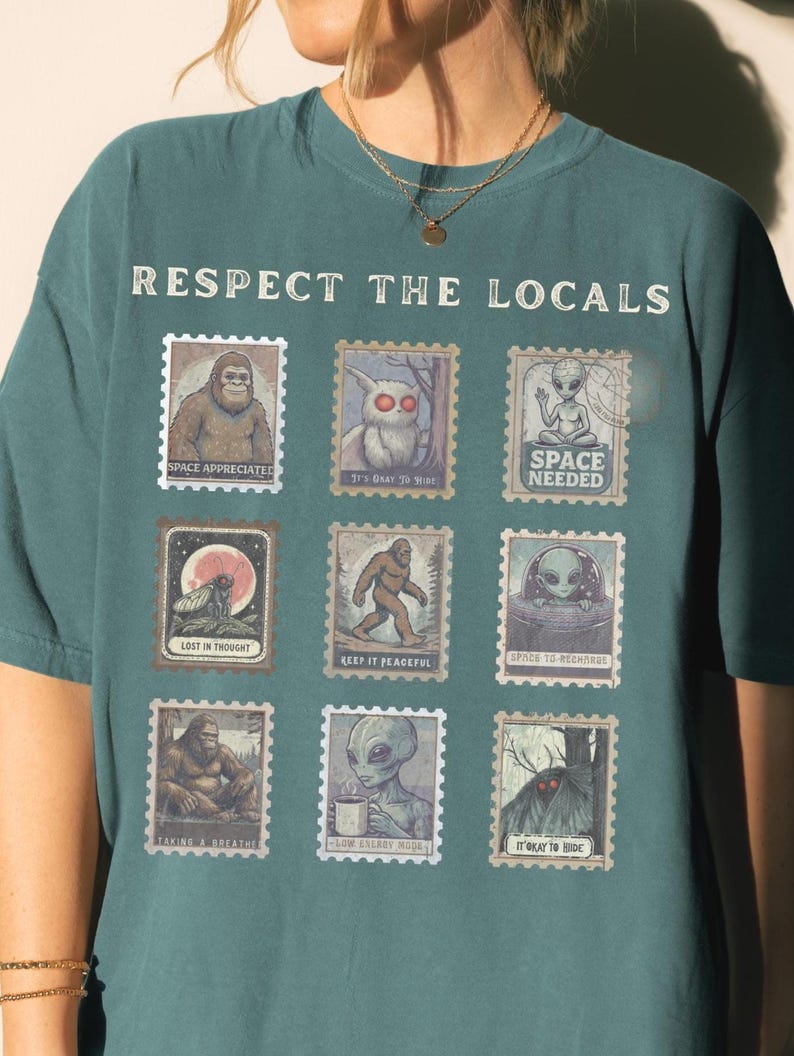 A woman wears a Blue Spruce colored Comfort Colors t-shirt with "Respect The Locals" cryptid stamp graphic, featuring Bigfoot, Mothman, and Alien stamps. Ideal for cryptid lovers and introverts.
