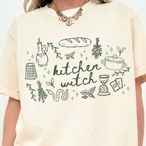 Woman wearing an Ivory Comfort Colors t-shirt featuring a kitchen witch design with bread loaf, herbs, cooking pot, olive oil bottle, tarot cards, snake, and botanical illustrations in a cottagecore style.