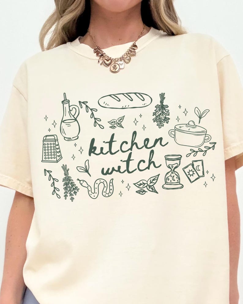 Woman wearing an Ivory Comfort Colors t-shirt featuring a kitchen witch design with bread loaf, herbs, cooking pot, olive oil bottle, tarot cards, snake, and botanical illustrations in a cottagecore style.