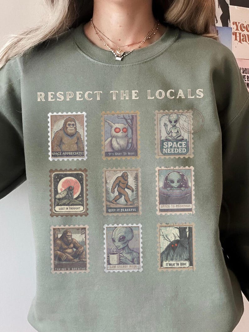Cozy cryptid sweatshirt with a grid of vintage postage stamp illustrations including Mothman, Bigfoot, and alien folklore characters shown on a model wearing a Gildan 18000 Military Green crewneck.