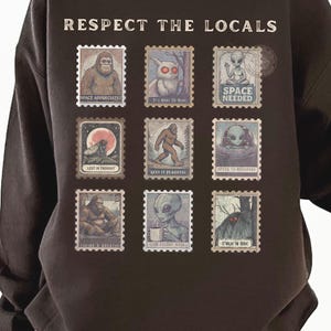 Cozy cryptid sweatshirt with a grid of vintage postage stamp illustrations including Mothman, Bigfoot, and alien folklore characters shown on a model wearing a Gildan 18000 Dark Chocolate crewneck.